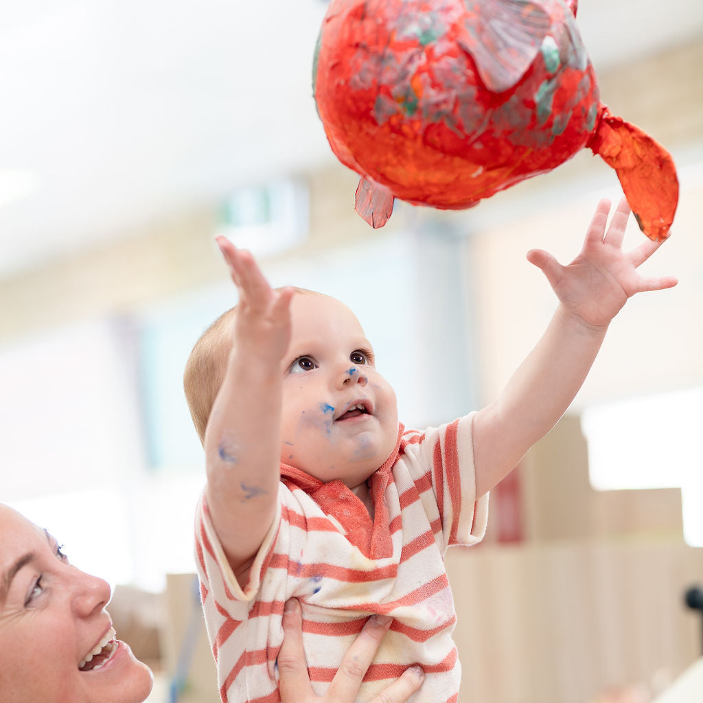 Baby reaching for a red ball held by an adult in a bright indoor setting