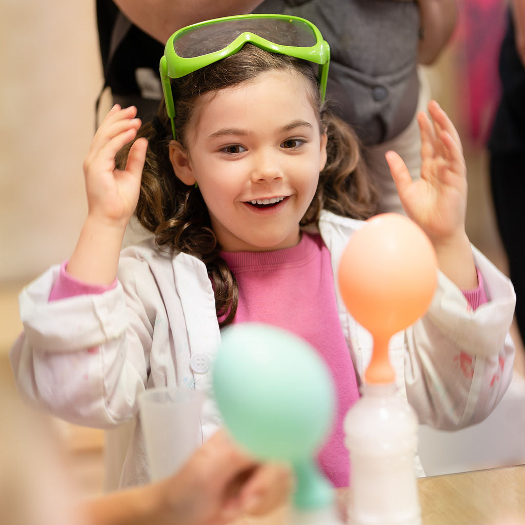 Child wearing green goggles participating in a science experiment with colorful balloons.